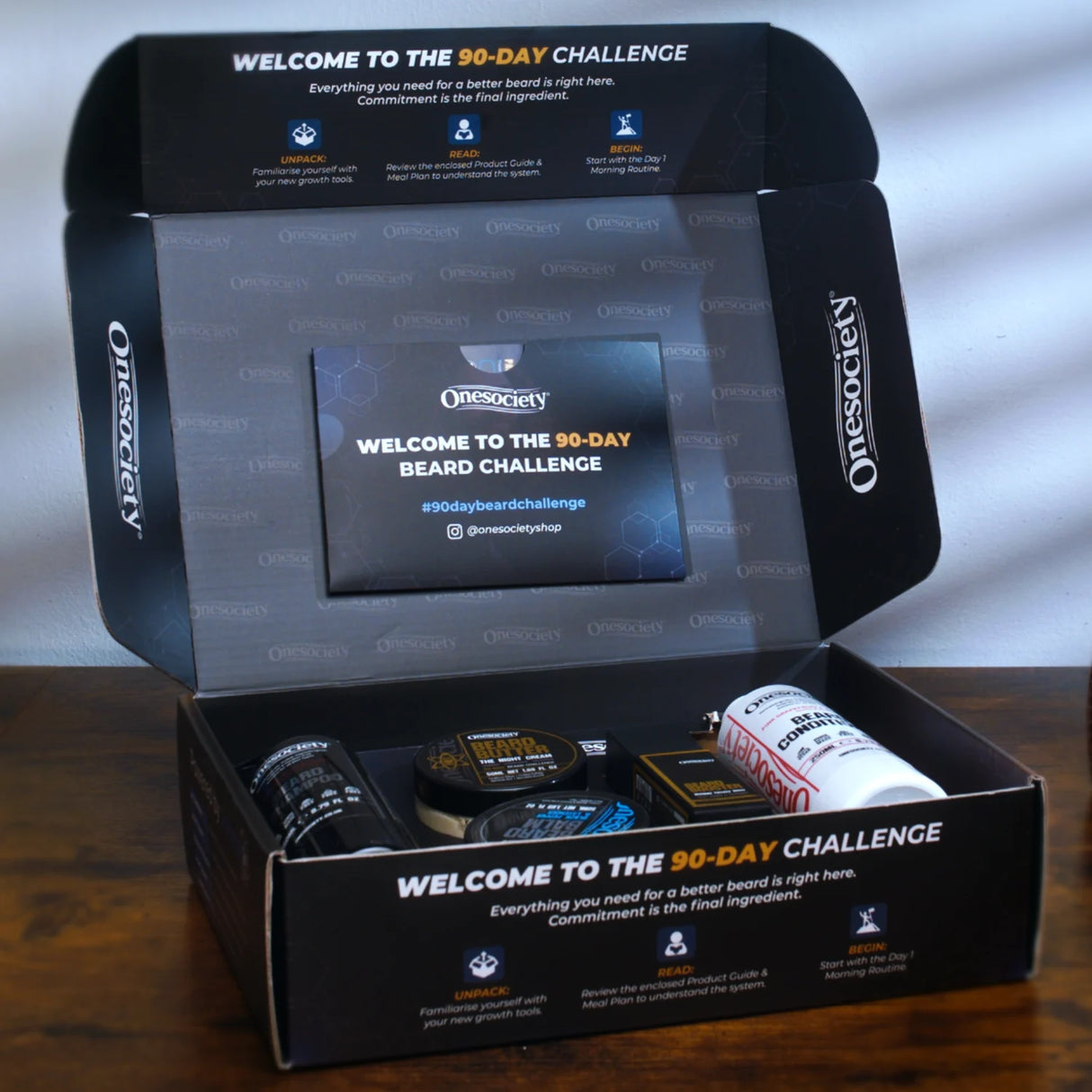 An open OneSociety 90-day beard challenge kit box on a wooden surface, showing the black branded packaging and several beard care products inside, including a beard butter, beard balm, ebard shampoo and conditioner and beard growth oil