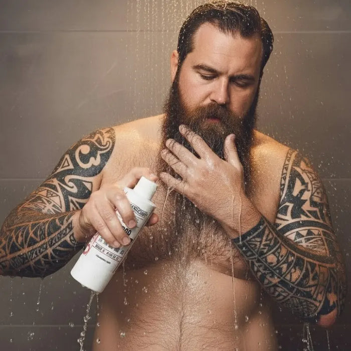 Man with tattoos applying a one society beard conditioner to his beard under a shower.