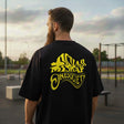 Man wearing a black t-shirt with yellow graphics and text, standing outdoors.