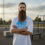 Man with a long beard wearing a white t-shirt with blue graphics in an outdoor setting.