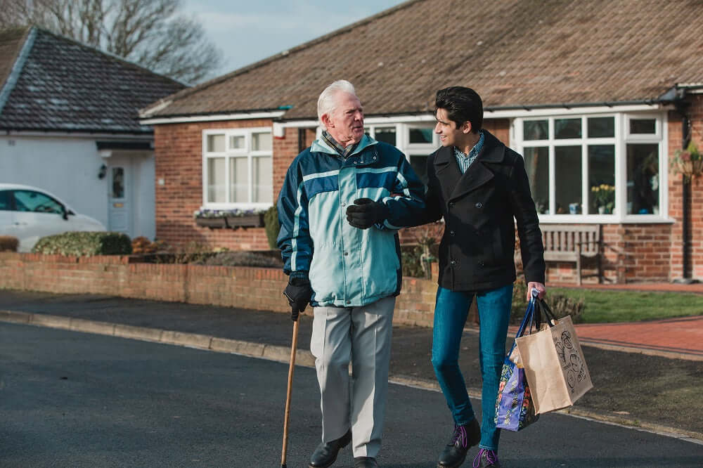 Young man heling old man with a walking stick cross the road and holding his shopping.